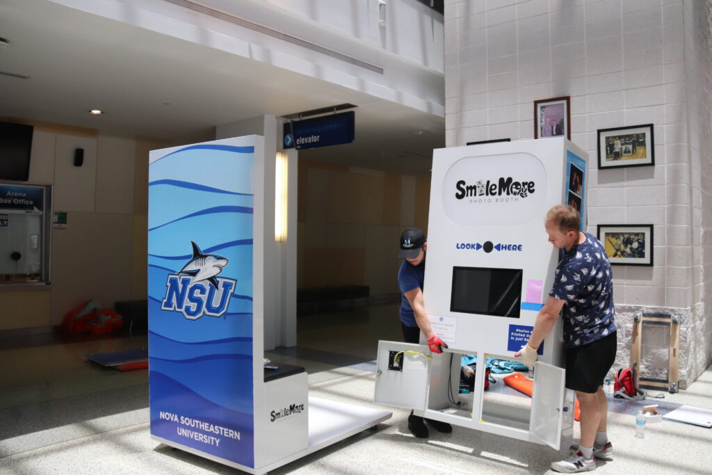 Two installers assembling a co-branded SmileMore photo booth in the Nova Southeastern University Center lobby